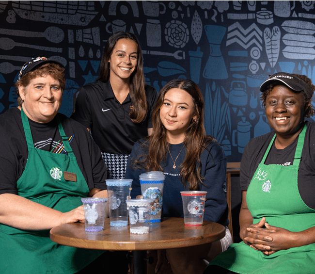 Lorena sits at a bistro table, three other women around her including two wearing green Starbucks aprons.