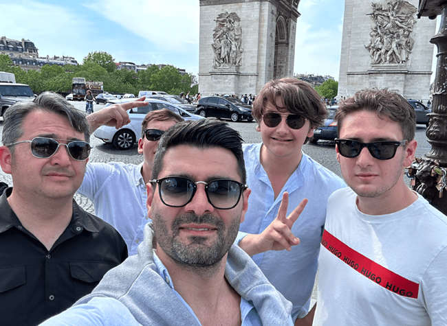 Sam holds out a phone for a selfie with four other men, the Arc de Triomphe in the background.