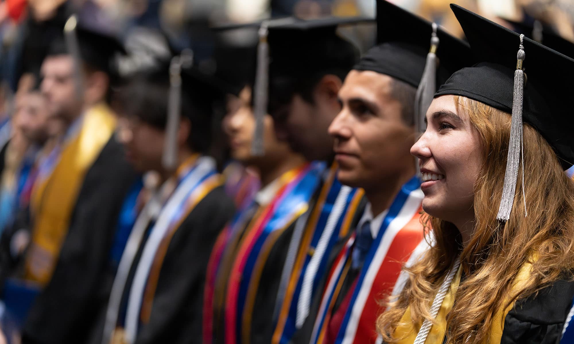 A line of smiling graduates wearing their caps, gowns, and various colors of sashes and cords.