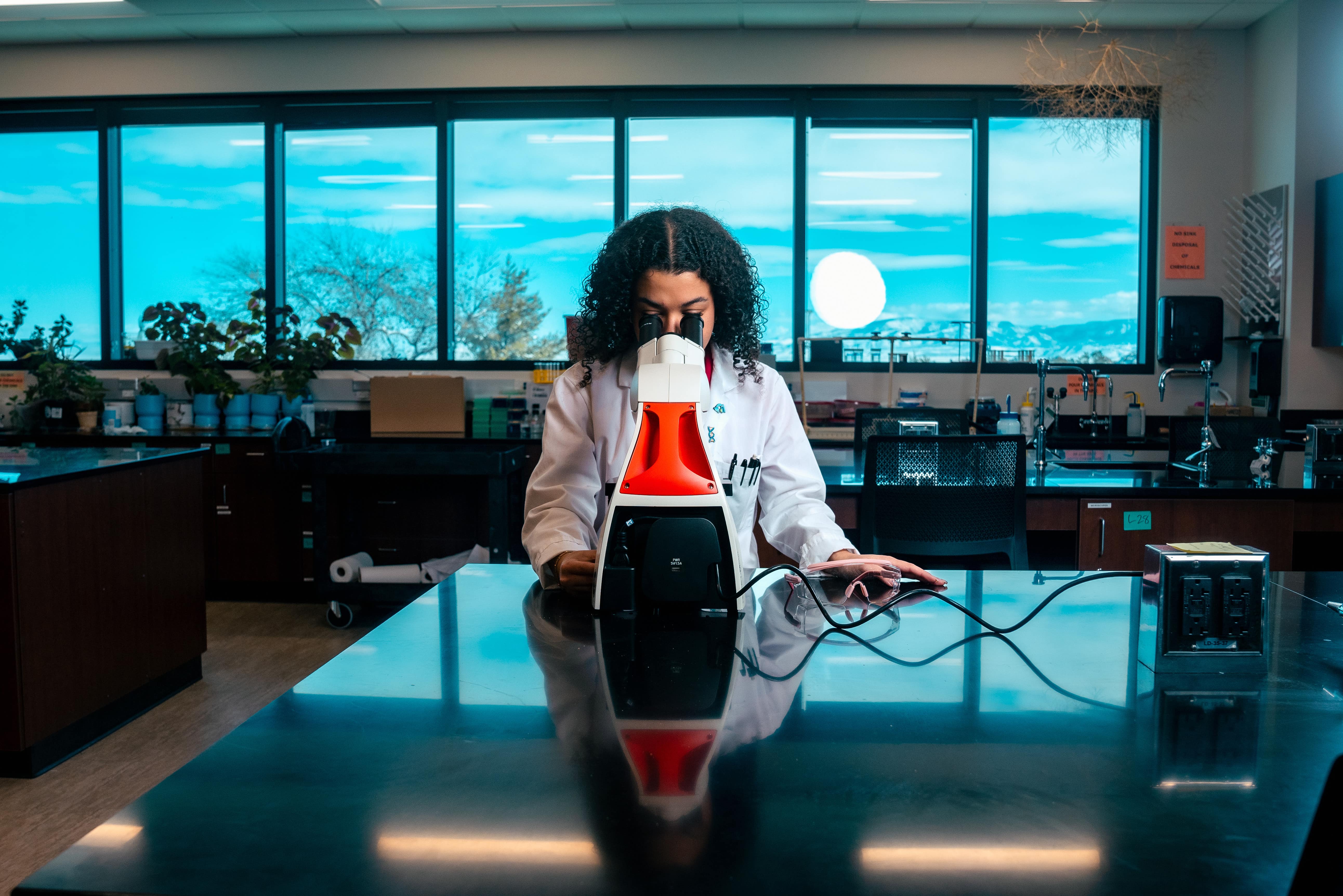 A student wears a lab coat and looks into a microscope. Mountains can be seen through the wall of windows in the background.