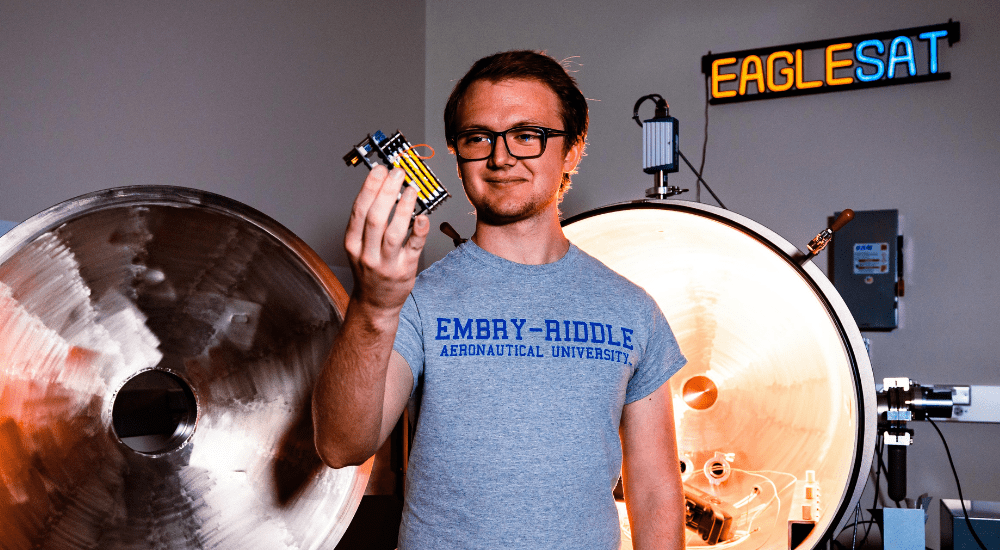 Hayden West in front of the thermal vacuum chamber holding a prototype electrical power system used to subject electronics to a space-like environment.
