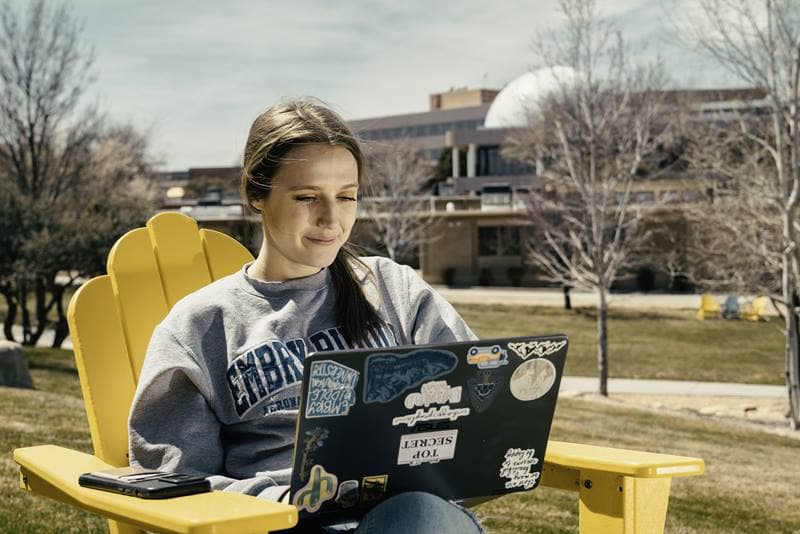 Emma sits in a yellow deck chair with a laptop in her lap, outside on the Prescott Campus.