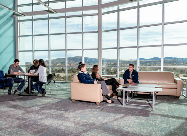 Several students are seated on couches in front of a large bank of windows through which low mountains are visible.