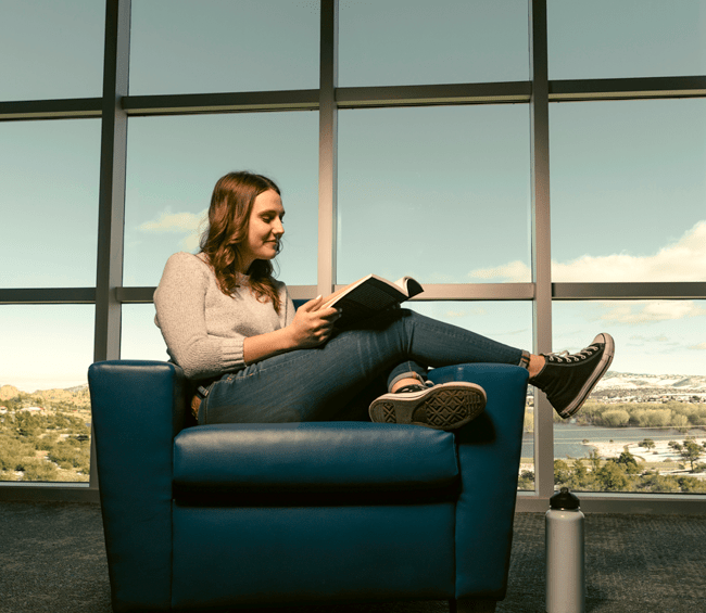 Emma lounges on an arm chair, reading in front of a bank of windows.