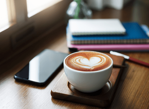 A latte, with a heart traced into the foam, sits on a desk next to a phone and a stack of books.