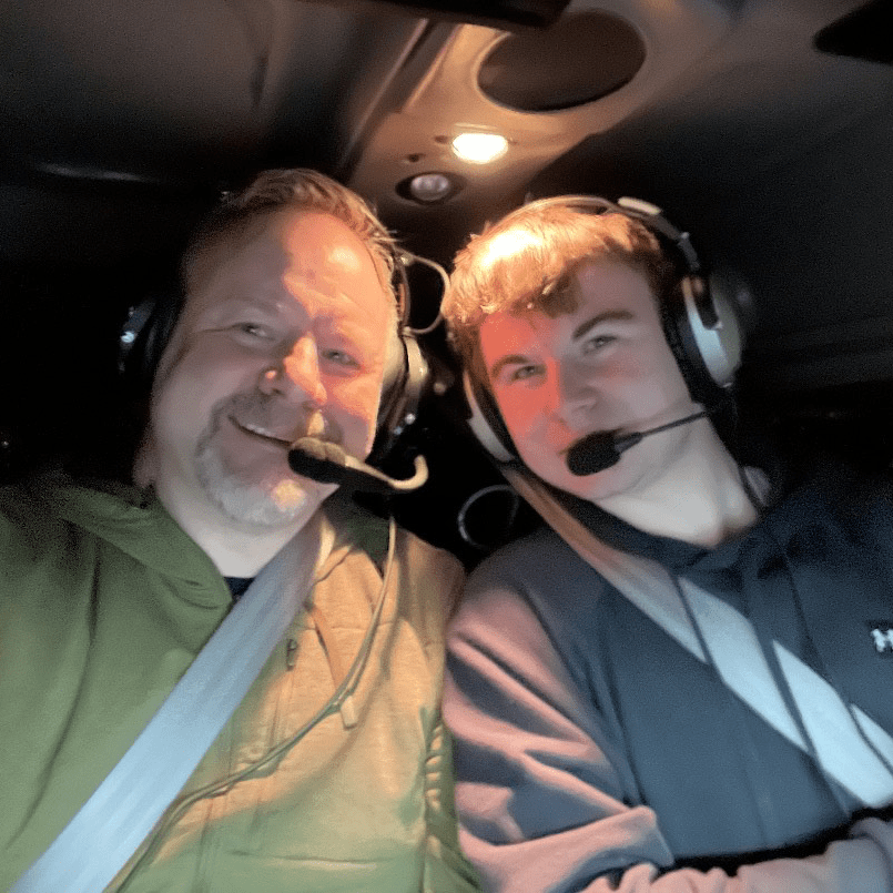 Killian and Paul wearing flight headsets and seatbelts lean together to pose for a selfie inside a plane.