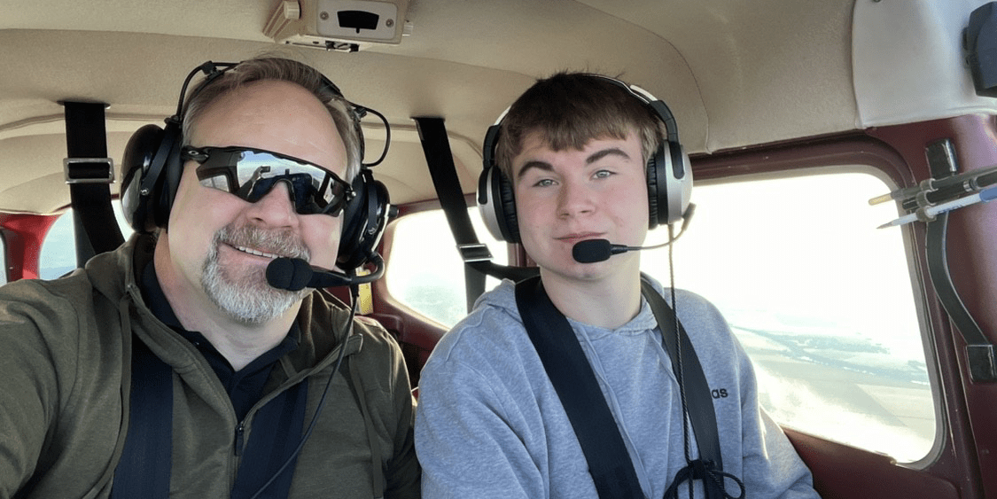 Killian and his father, both wearing flight headsets, pose in the cockpit of a plane.