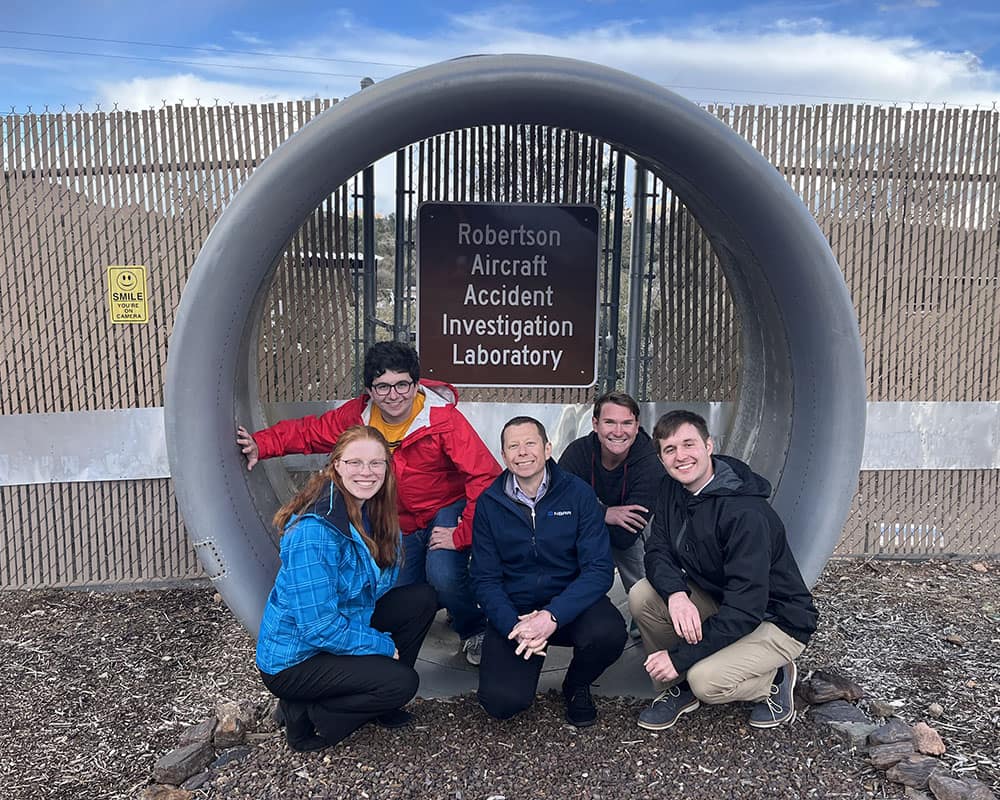 Alex poses with four other students, crouched inside an empty engine pod, in front of a sign on a fence that reads, Robertson Aircraft Accident Investigation Laboratory.