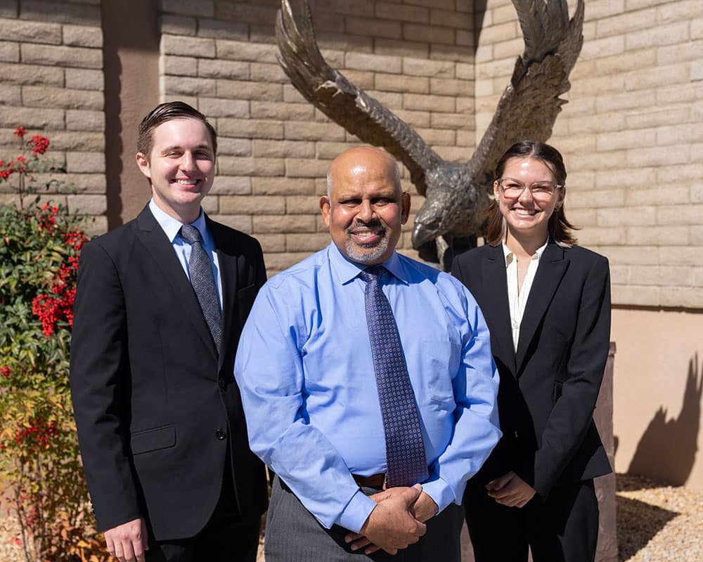 Three people in business wear pose outside in front of a flowering bush and an eagle sculpture.