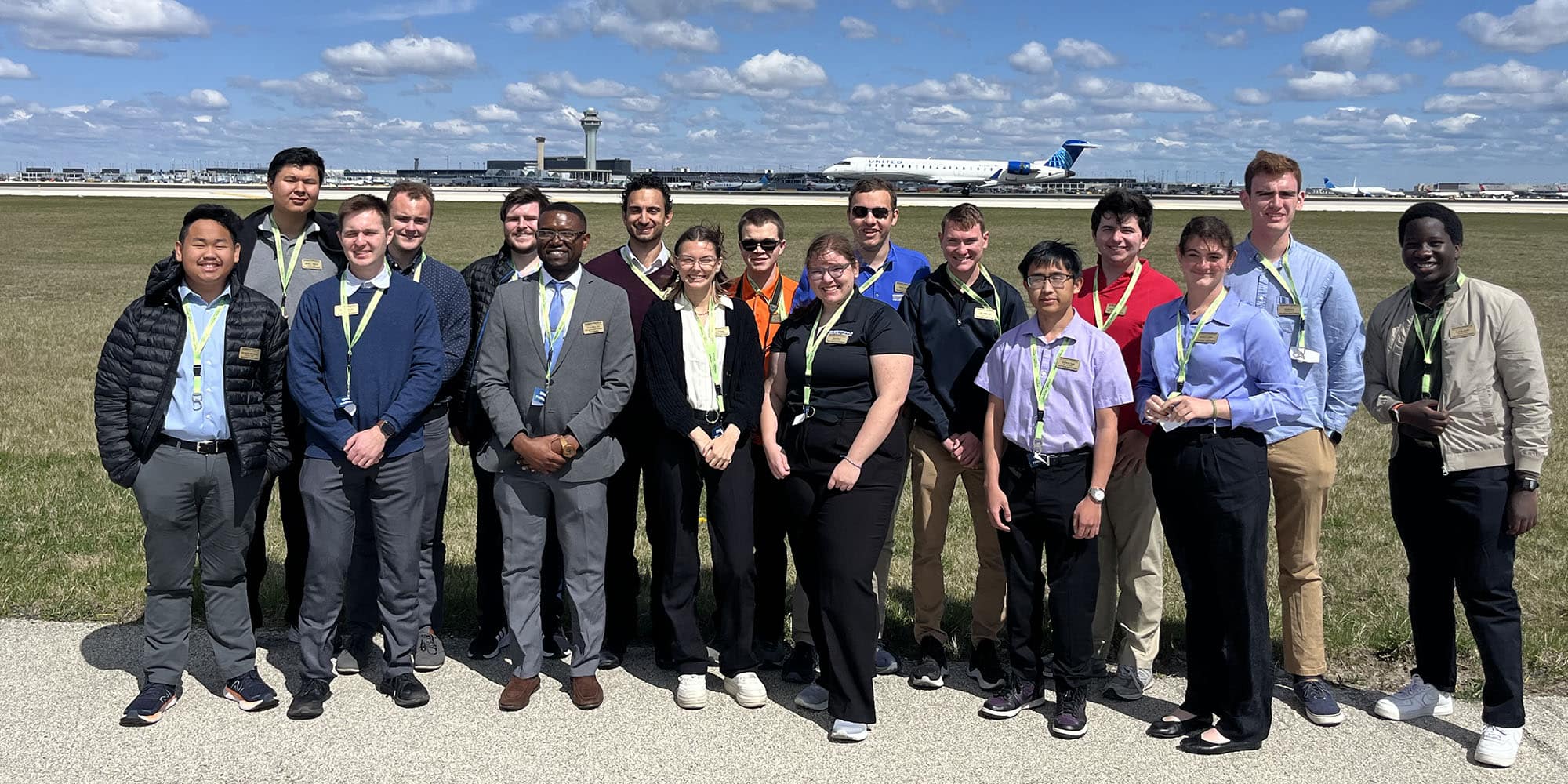 Seventeen students in business wear, with yellow lanyards, pose in the sun in front of a tarmac, a flight control tower in the distance.