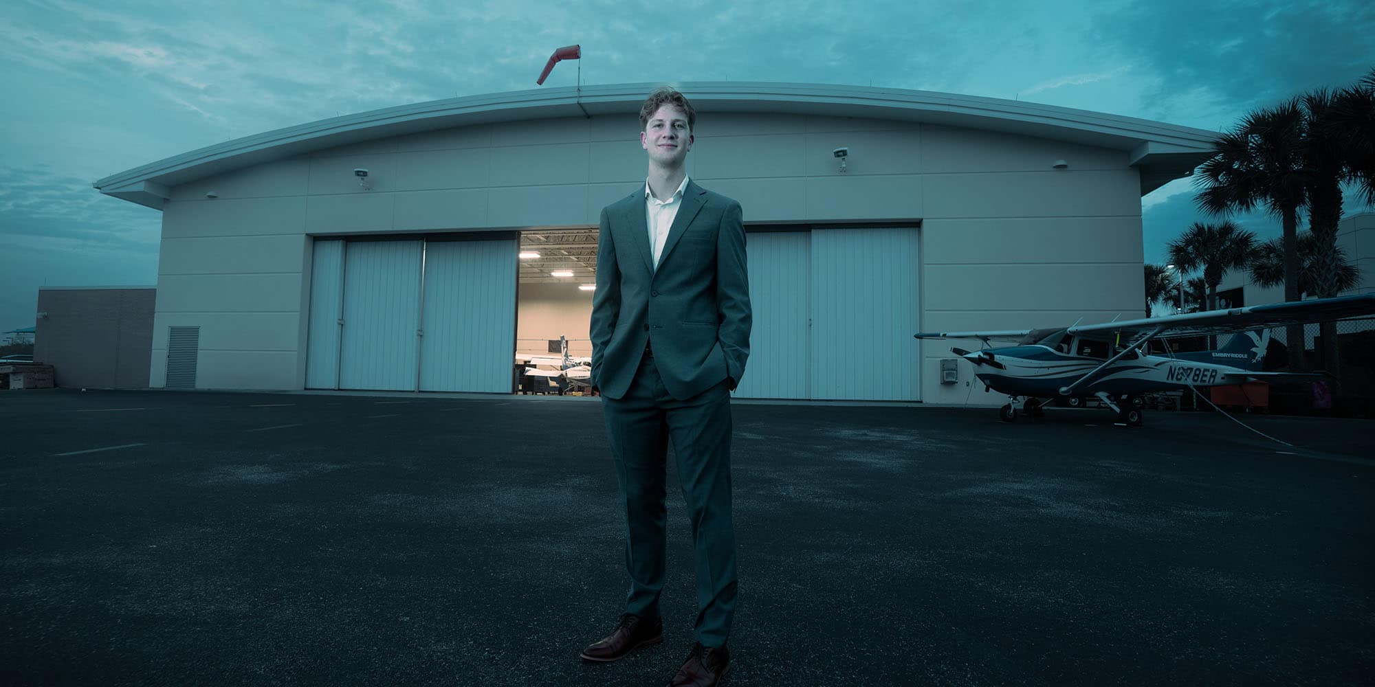 Arthur wears a business suit and no tie, posing in front of a hangar at dusk.