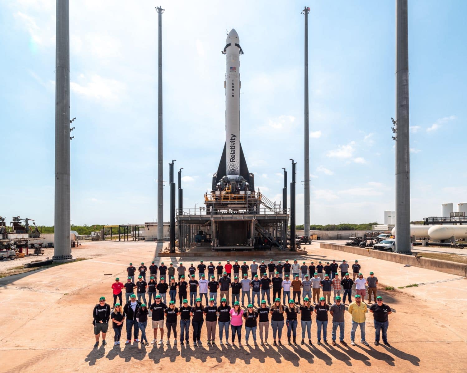 A wide shot of three rows of twenty people in bright green baseball caps, standing in front of a rocket on a launch pad.