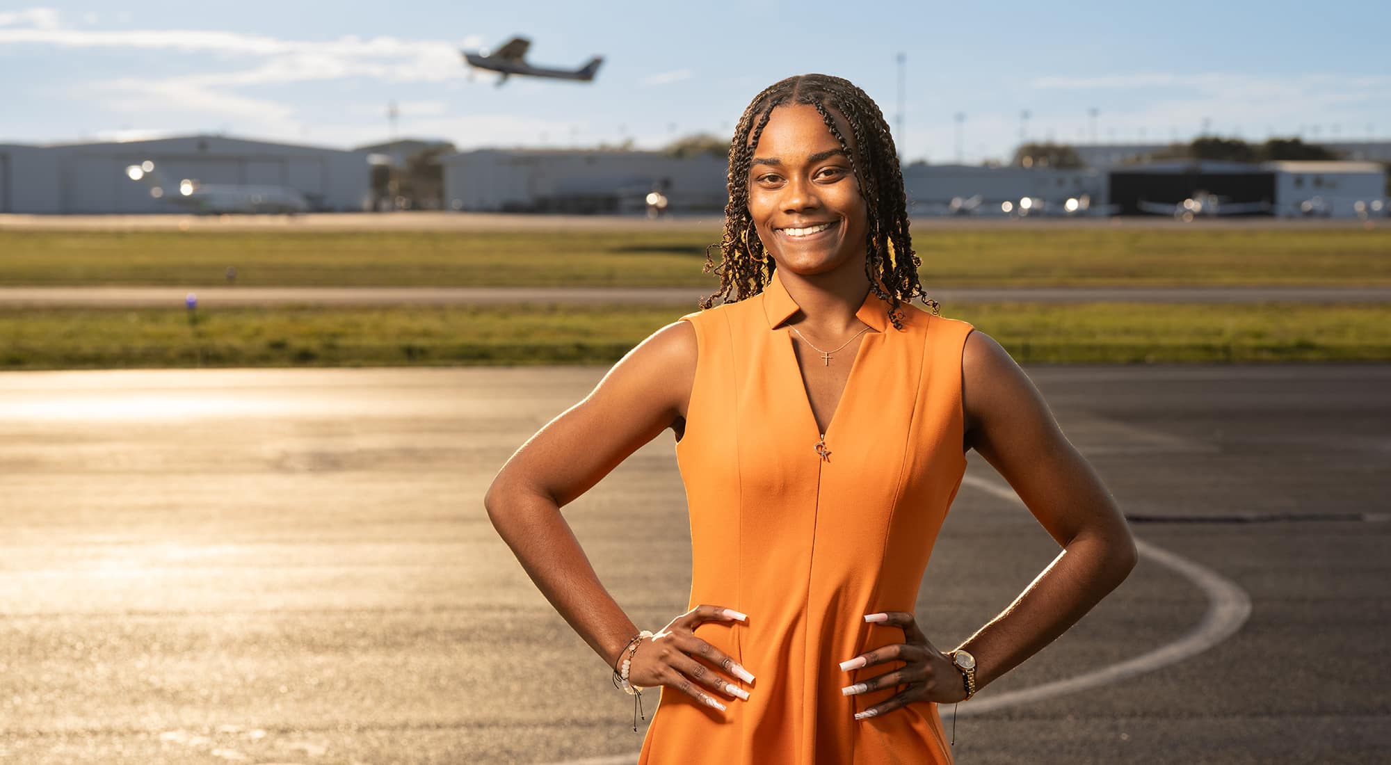 Coral, with long braided hair, poses in an orange dress, a small plane taking off in the distance.