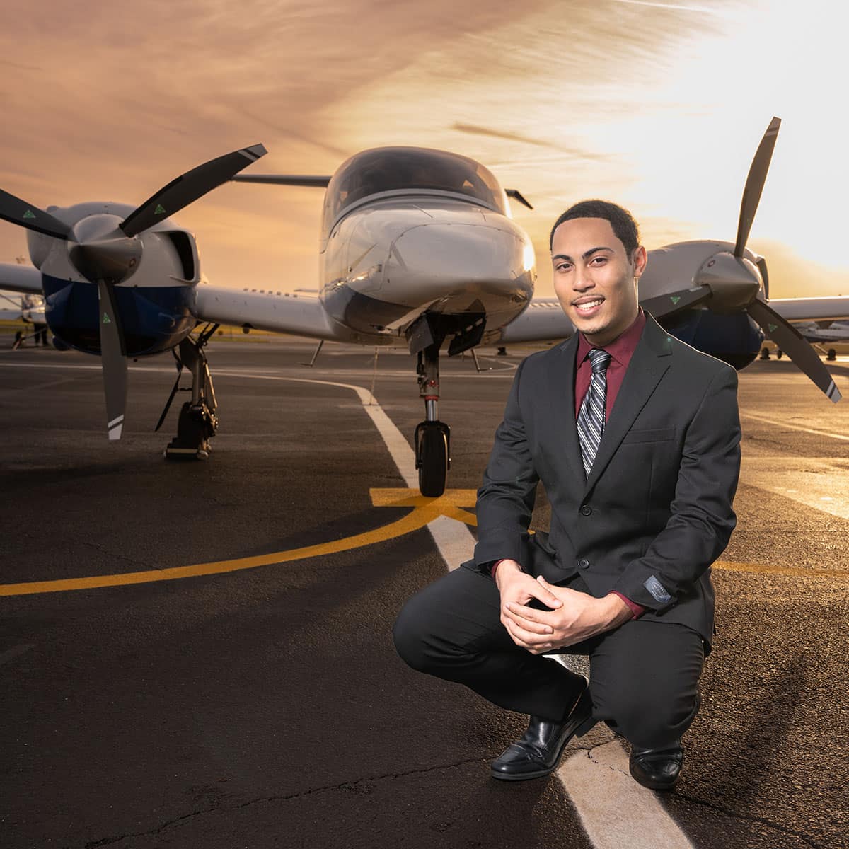 Dariel, wearing a black business suit and tie, crouches in front of an aircraft at sunset.