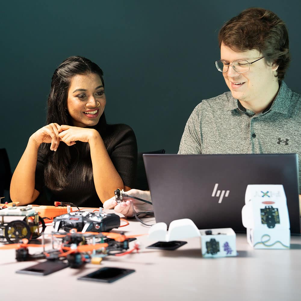 Two people sit at a table in front of an open laptop and drone parts.