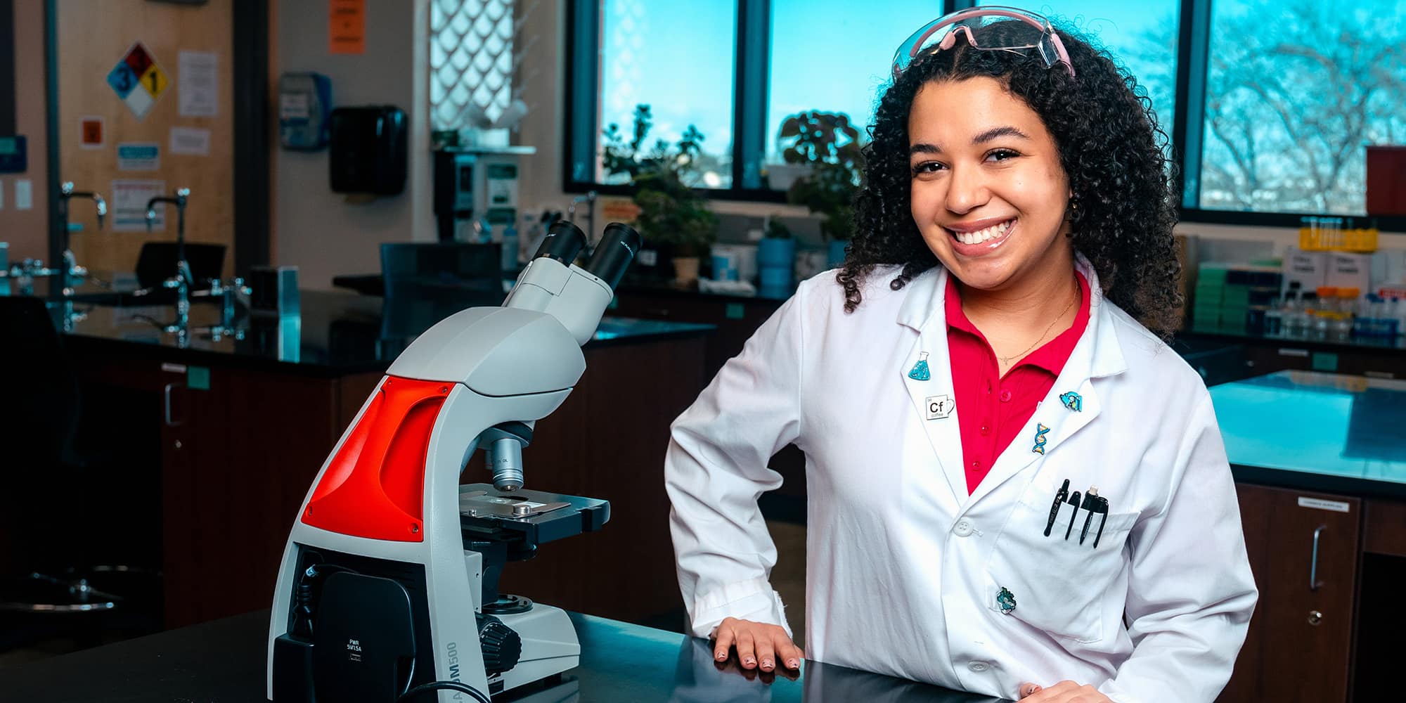 Makenzi, with curly hair, smiles in a white lab coat next to a microscope.
