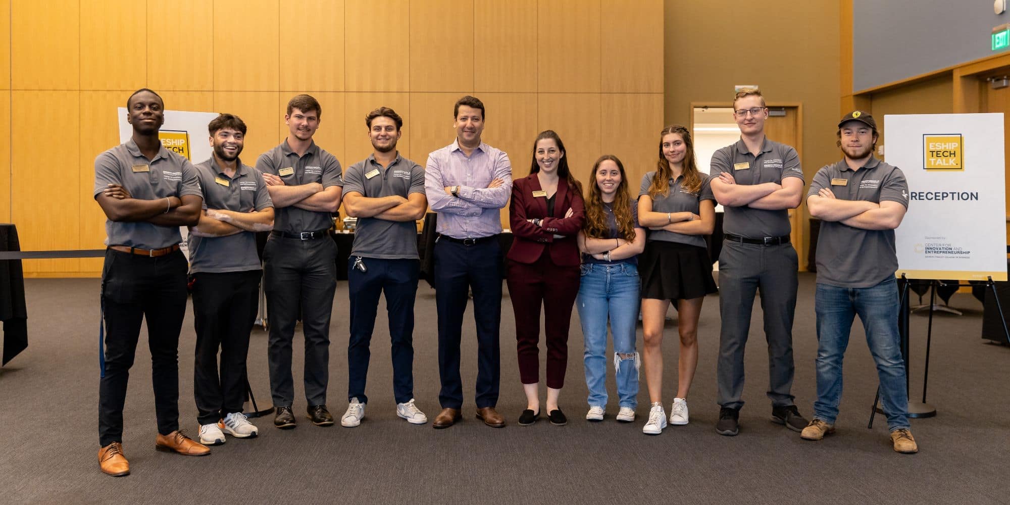 Eights students in grey Embry-Riddle polos pose with instructors in business wear next to a sign on an easel that reads E SHIP Tech Talk Reception.