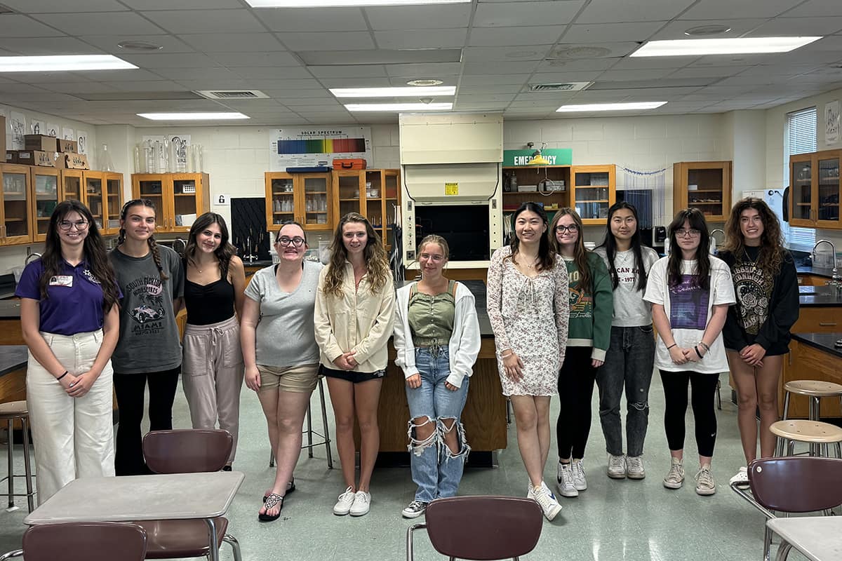 Eleven women casually dressed line up in front of a classroom with laboratory equipment.