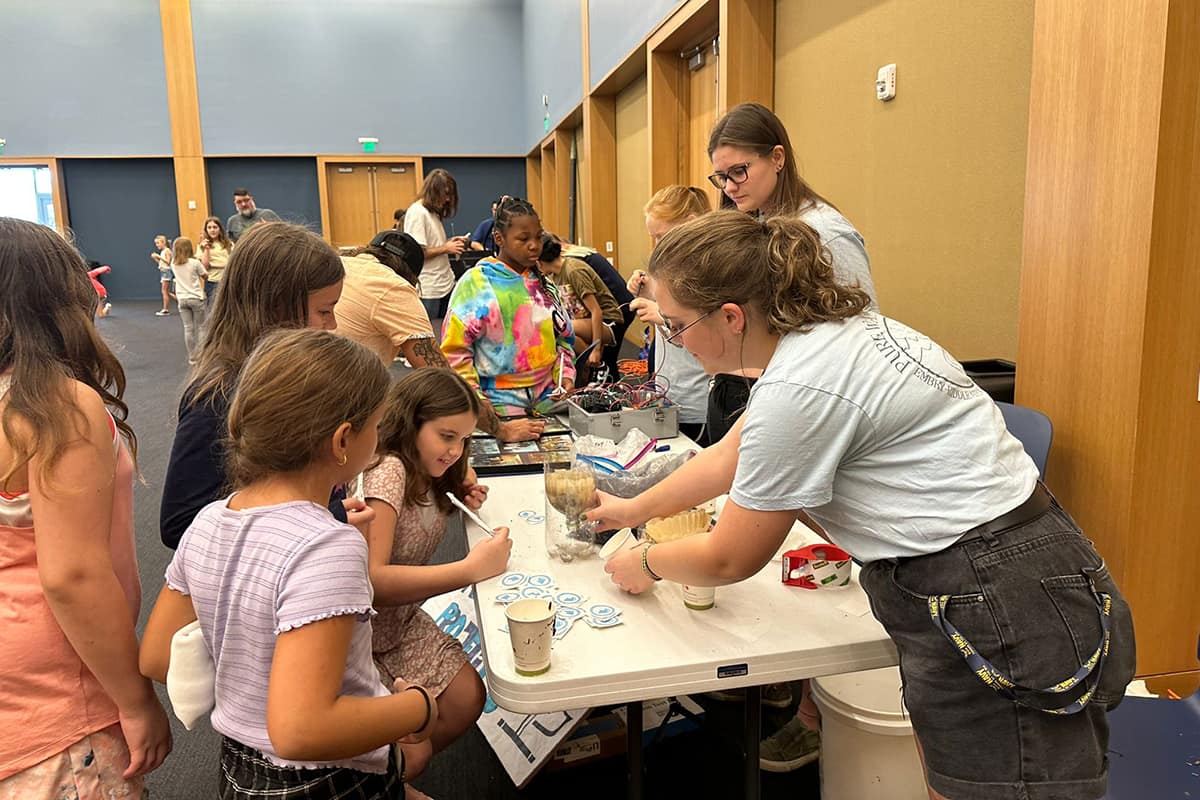 Students help young girls with projects at several tables in a conference hall.