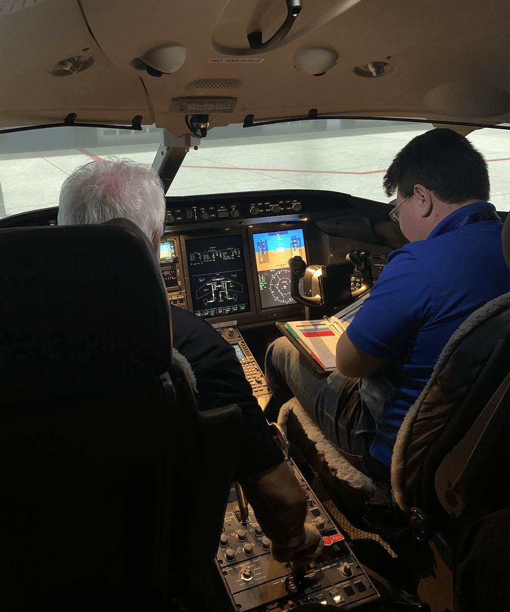 Timur studies a manual, seated at the controls of an aircraft alongside an instructor.