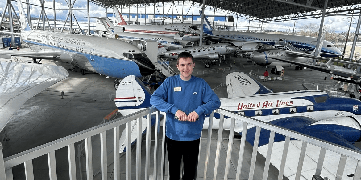 Timothy leans against a railing in front of several jets parked under a large canopy.