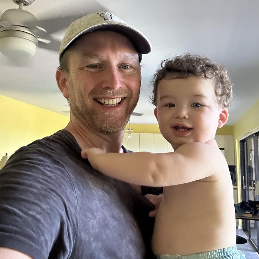 David takes a selfie, holding a small child, in a home setting.